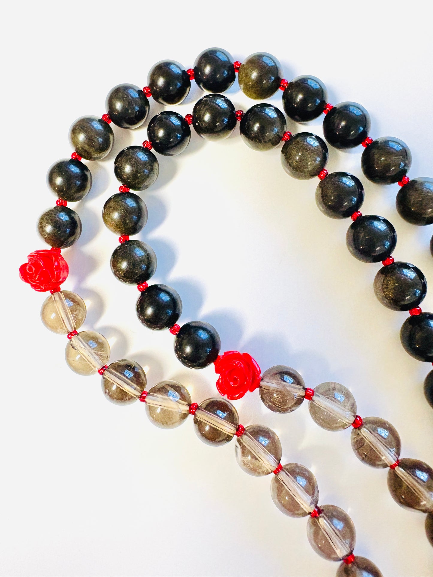 Close up of mala necklace with golden sheen obsidian, smoky quartz, and red rose beads on a white background